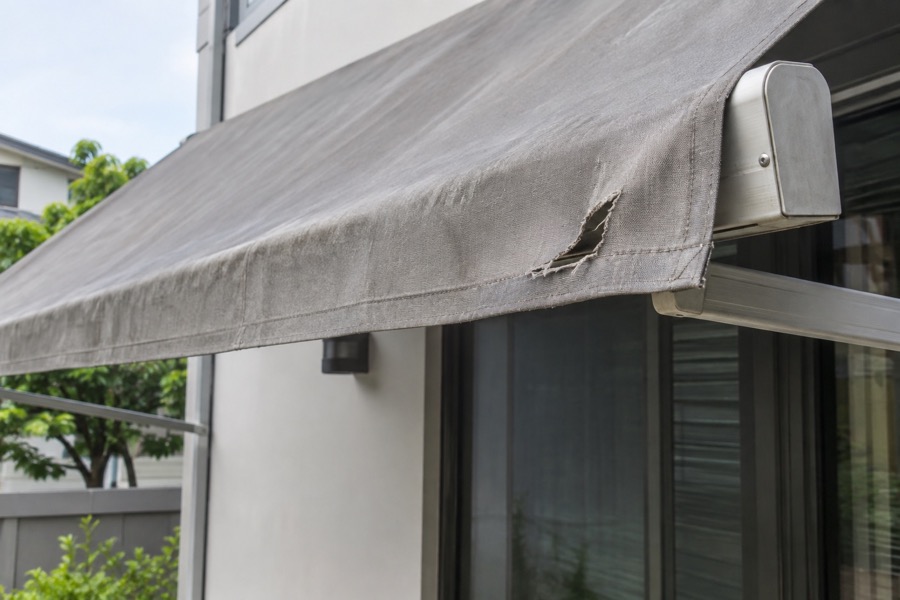 Close-up of a small tear near the edge of an aged grey awning fabric stretched on its intact frame, clearly visible repair concern