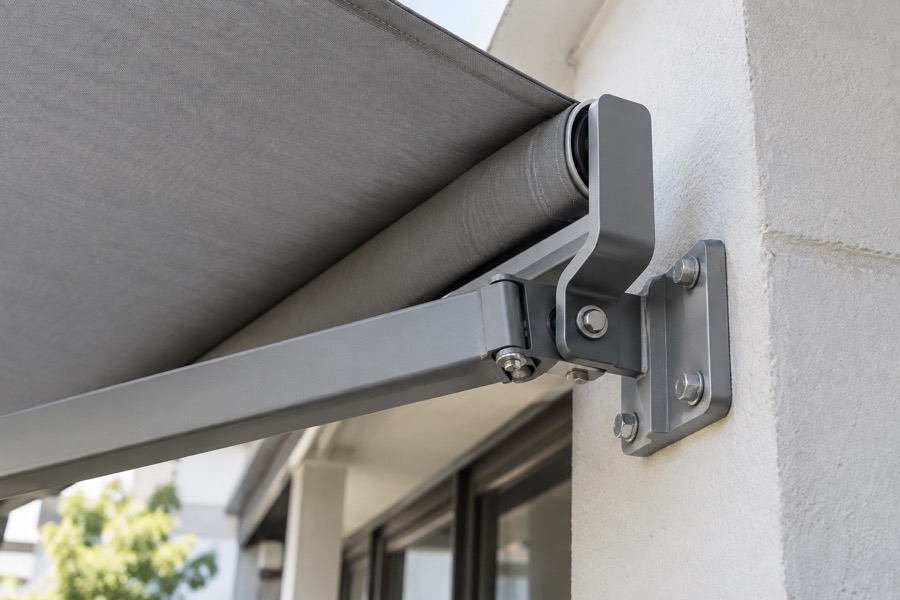 Close-up of a correctly mounted grey folding-arm awning wall bracket on a painted concrete wall, arm joints and bolts visible