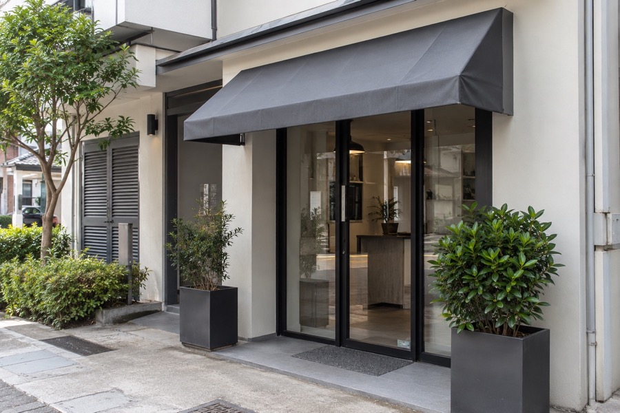 Small commercial entrance with a compact grey fixed fabric canopy and clear pedestrian pavement, simple utilitarian facade