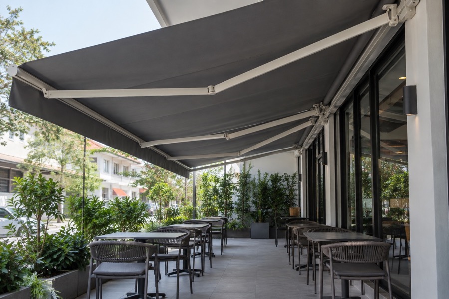 Underside view of folding-arm awning over an empty outdoor café seating area, plausible arms and hardware, focus on clear floor space