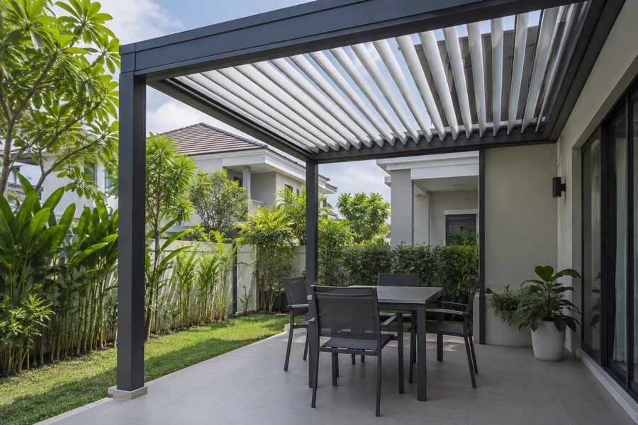 Modest residential patio beneath an adjustable aluminium louvre roof, slats partly open, simple powder-coated frame and empty table