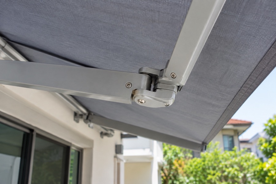 Close-up of a folding-arm joint beneath a taut grey awning canopy, neat metal finish, credible hardware