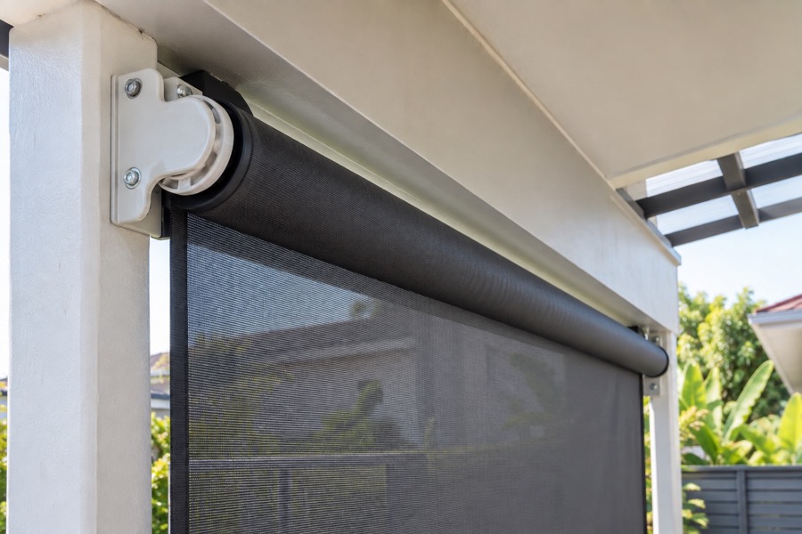 Close-up of outdoor roller-blind top roller and wall brackets underneath a covered verandah beam, mechanically plausible