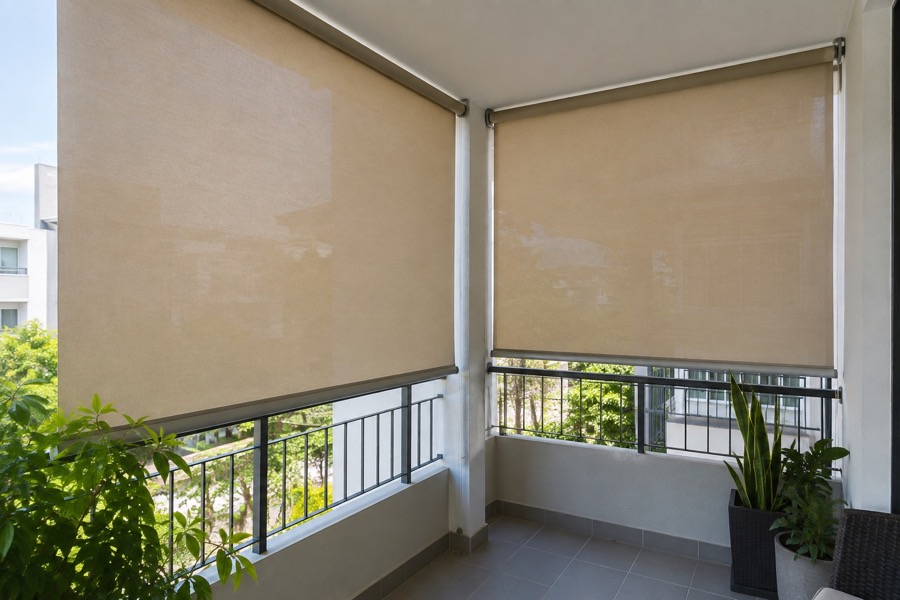 Two lowered beige outdoor roller shades screening a small residential balcony side opening, simple tiled floor and plants